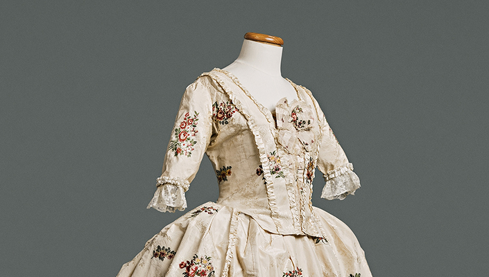 Une robe à l’anglaise sur un mannequin. Le corsage, ajusté devant et dos, présente une taille marquée par une couture. De couleur crème, elle est ornée de fleurs brodées et de poignets en dentelle à volants.
