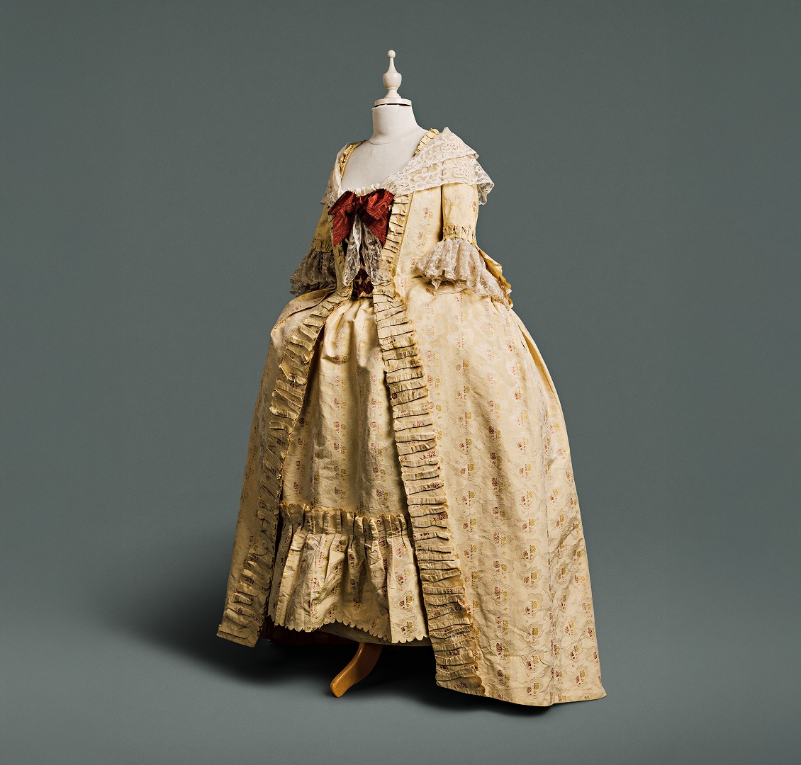 Une robe couleur crème, ornée d’un empiècement fluide au dos du corsage, dévoilant un tissu précieux à motifs de grenades, de roses et autres fleurs, sur un fond de dentelle damassée. Un grand nœud auburn et un châle de dentelle drapent le haut de la robe.