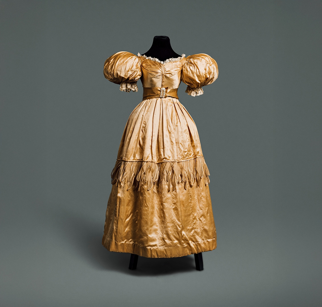 Robe en soie dorée avec une longue jupe ample, des manches bouffantes autour des épaules et une taille fine soulignée par une ceinture de même matière.