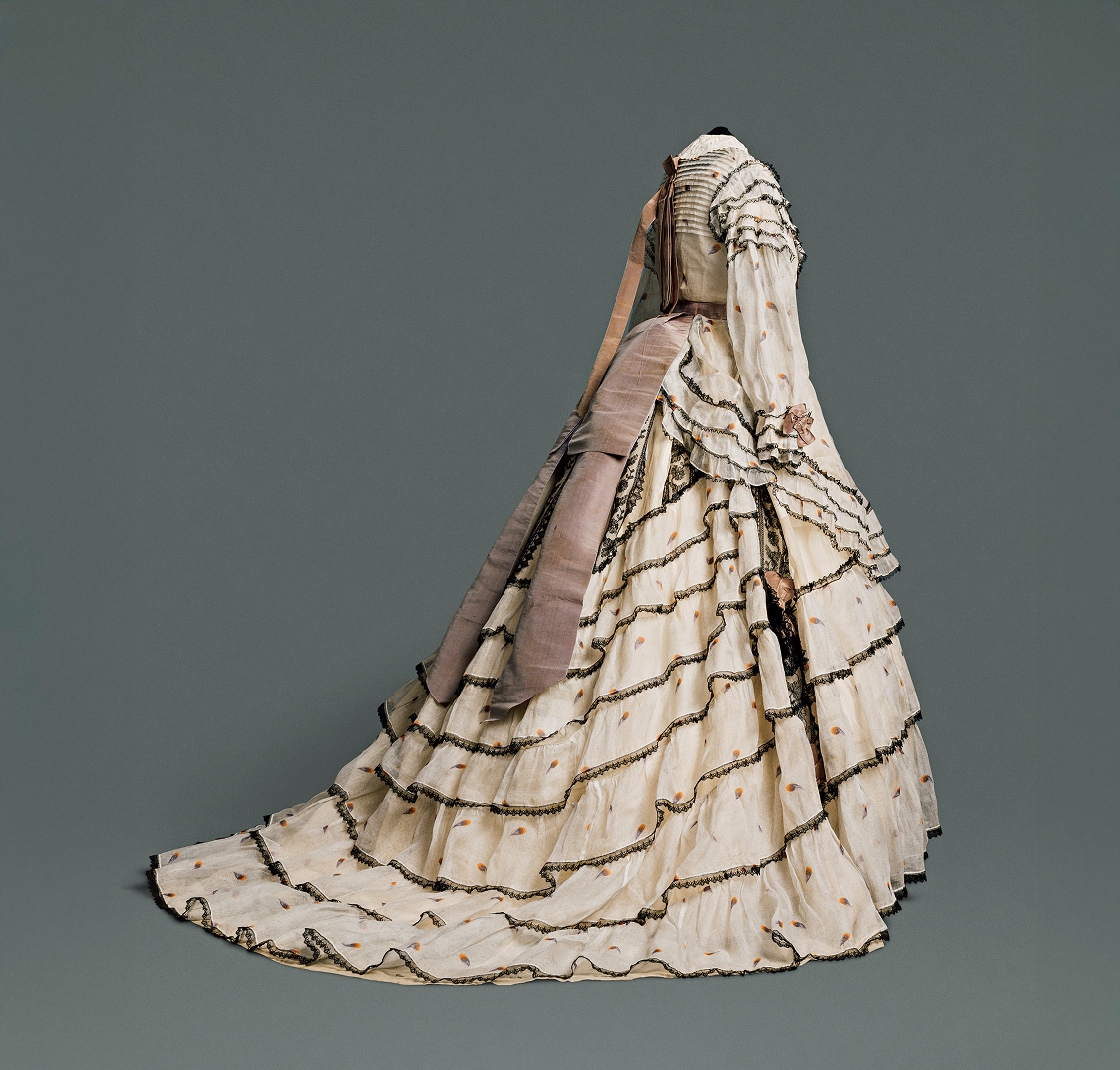 Une longue jupe volumineuse composée de plusieurs couches de coton et de lin à motifs couleur crème, rehaussée de dentelle noire. Une ceinture marron clair descend de la taille, à l’arrière de la robe.