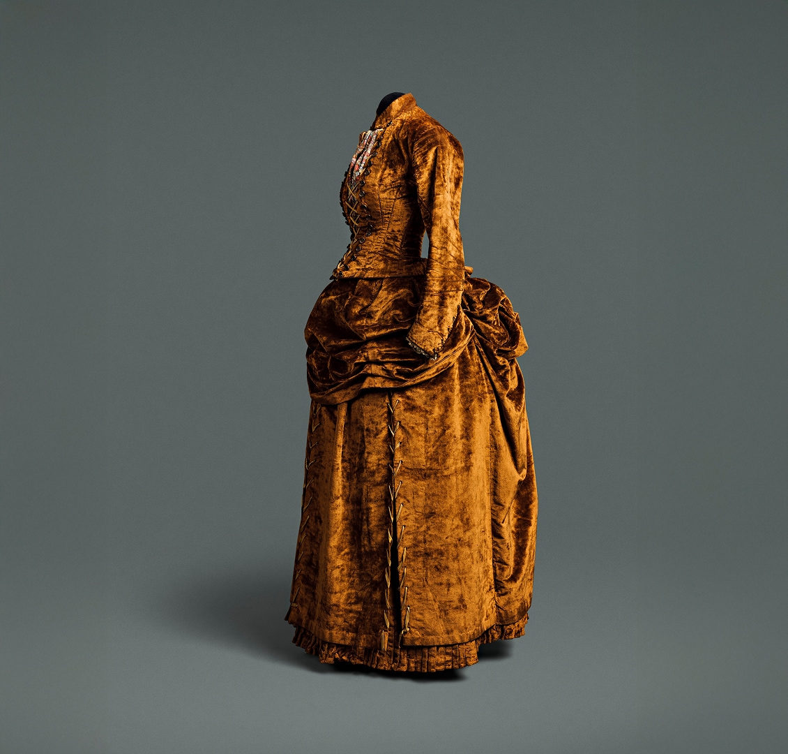 Une robe ocre foncé composée d’un corsage et d’une jupe séparés. La robe est ornée de boutons décoratifs marron qui descendent le long du corsage et sont lacés par un cordon doré.
