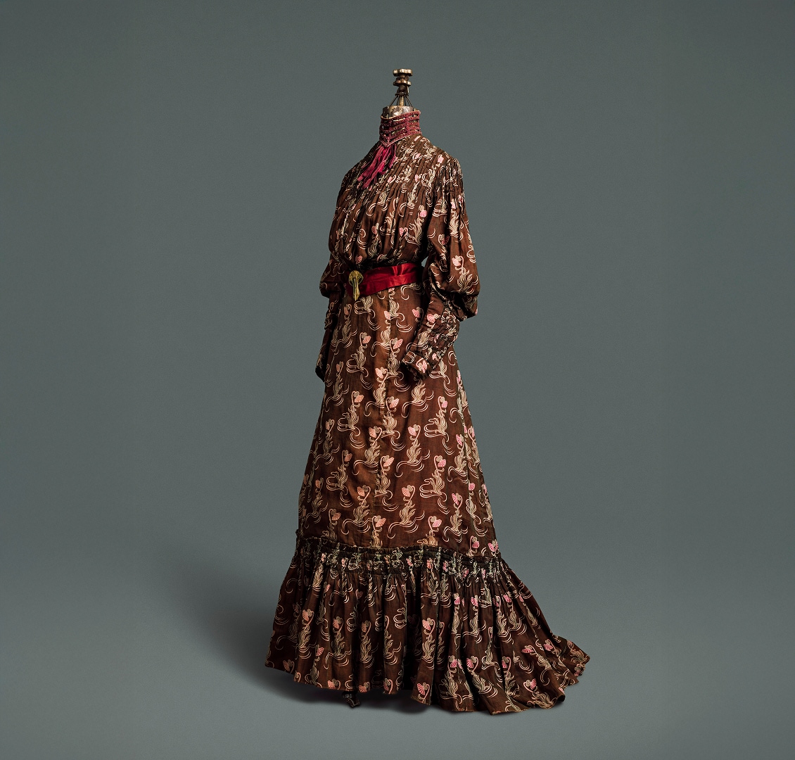 Une robe quatre pièces en coton marron chocolat à motifs de chardons marron clair et roses. La robe est rehaussée d’une ceinture en soie rouge foncé, fermée par une agrafe dorée, et d’un col.