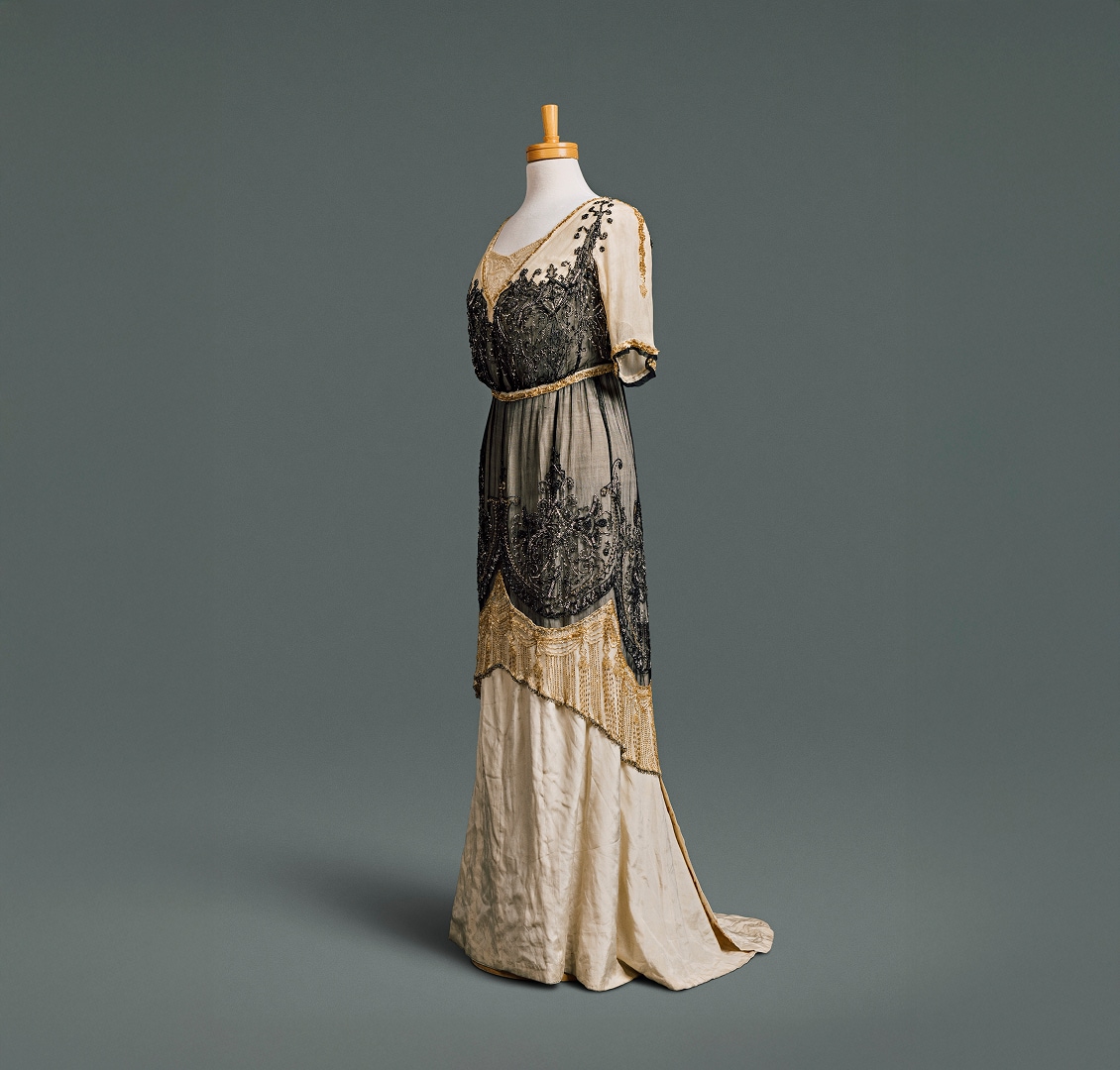 Une longue robe étroite en soie couleur crème, à décolleté plongeant et taille haute, recouverte d’une surjupe tunique ornée de perles noires et dorées.