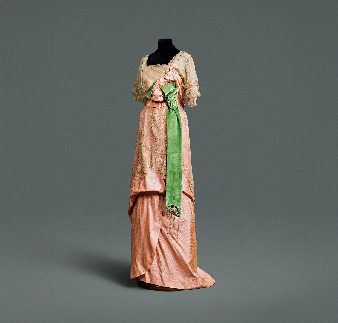 Une robe en soie rose à jupe drapée et large ceinture vert émeraude, recouverte d’un corsage et d’une surjupe en dentelle finement travaillée.