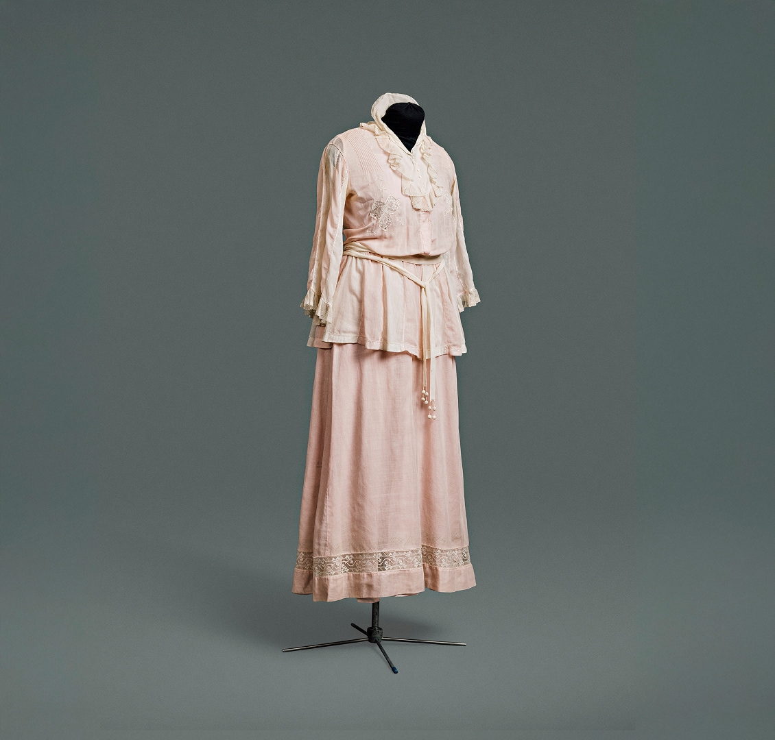 Une simple robe-robe rose en coton, conçue pour ressembler à un tailleur composé d’une veste sur un chemisier, portée avec une jupe assortie.
