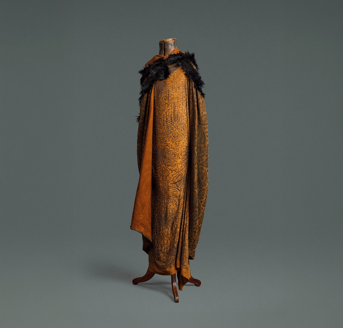 Une longue robe ample en soie auburn ornée d’un motif floral en perles noires. Elle est recouverte d’une cape assortie en tissu plus épais à motifs et d’un boa en plumes noires.