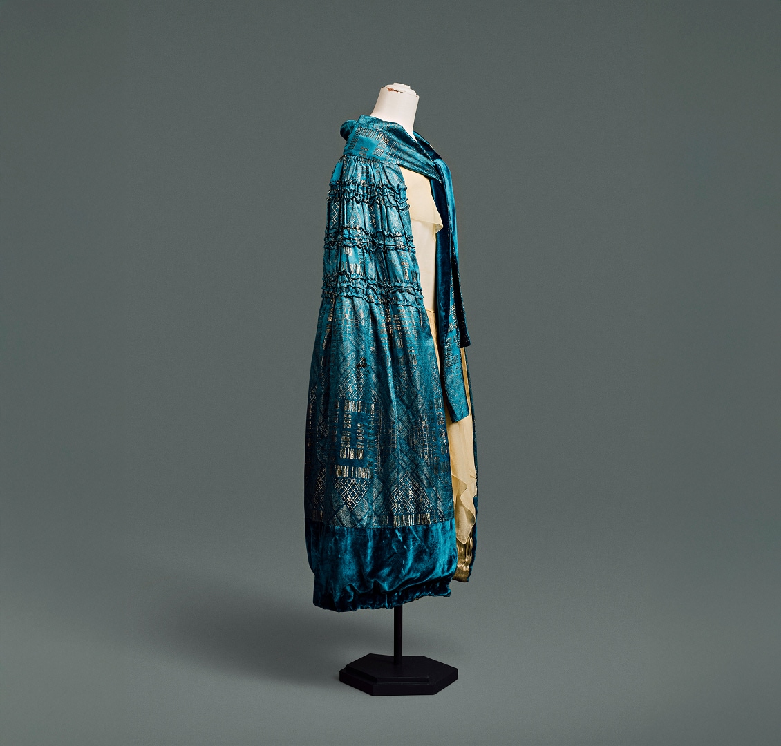 Une cape en soie bleue, ornée d’un motif damassé Art déco aux formes géométriques dorées métallisées, arrive juste en dessous du genou.