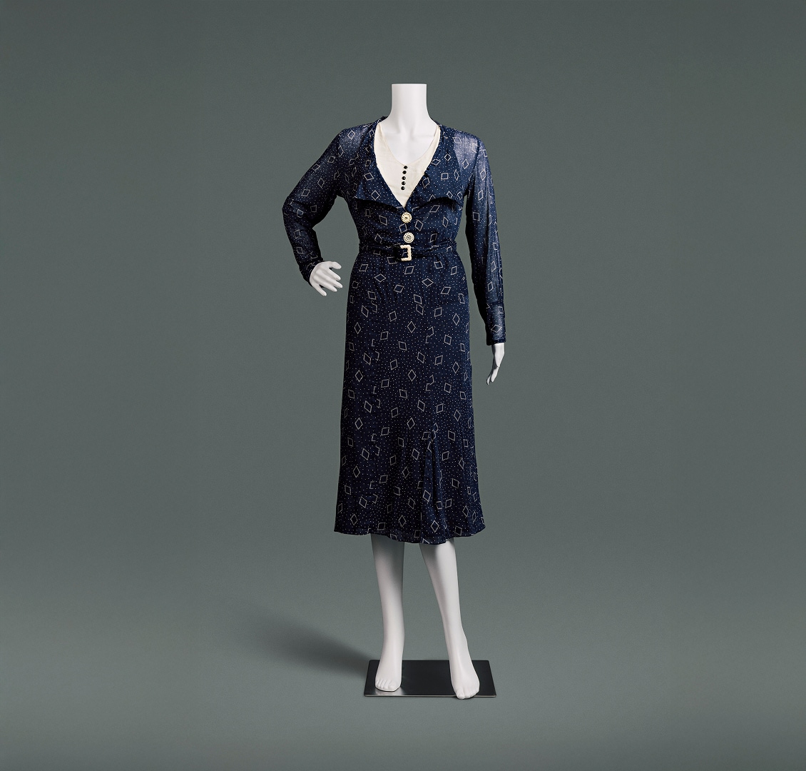Une robe en coton bleu nuit, cintrée, à taille haute et à jupe arrivant juste sous les genoux. Le tissu est orné de petits pois et d’un motif en losanges.