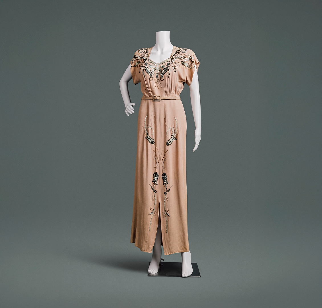 Une robe couleur pêche ornée de perles et de fleurs, aux épaules fortement rembourrées et à la jupe ajustée. Une fine ceinture du même tissu souligne la taille haute.