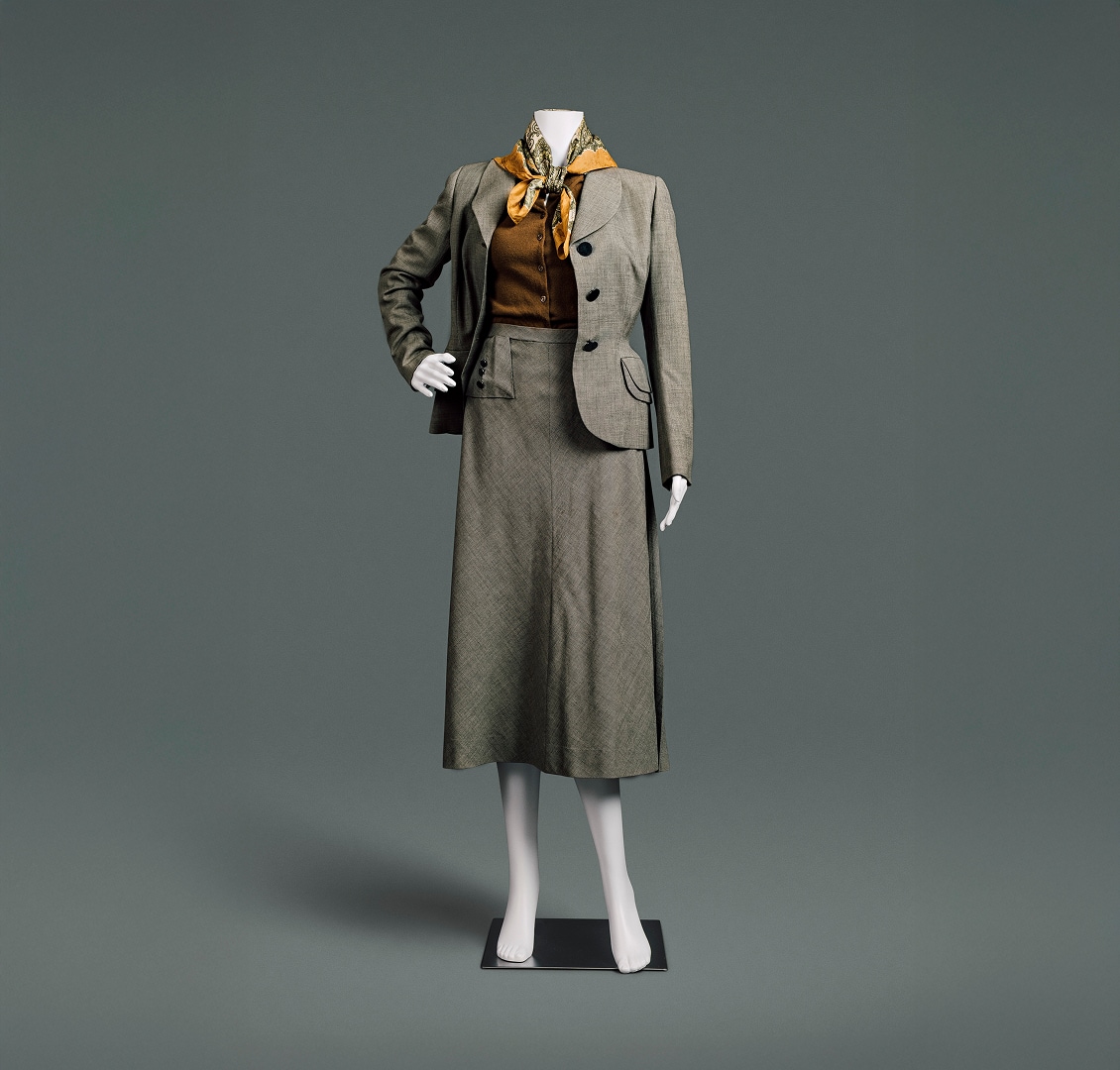 Un tailleur en laine grise anglaise, composé d’une veste et d’une jupe. Ce tailleur présente des épaules tombantes et larges, une blouse boutonnée auburn, une taille fine et une jupe longue et ample. Un foulard à motifs cachemire noué autour du cou apporte la touche finale parfaite.