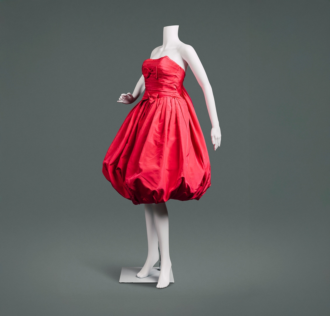 Une robe cocktail rouge cerise sans manches en soie, avec une jupe ballon, un dos sac et des nœuds rouges au niveau de la poitrine et de la taille.