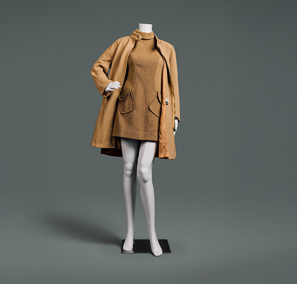 Une robe courte en laine couleur camel, à col roulé, à motif chevrons et poches en biais, accompagnée d’un épais manteau camel qui descend juste au-dessus du genou.