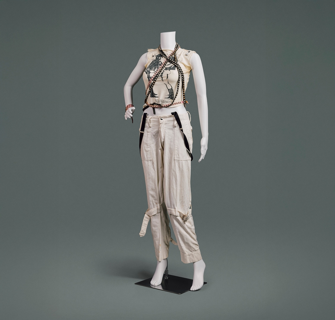 Un t-shirt à manches déchirées orné de l’emblématique imprimé « Two Cowboys » de Vivienne Westwood sur le devant, un pantalon en coton couleur crème avec de grandes poches et des ceintures cousues qui se resserrent autour de chaque jambe au-dessus du genou, et des ceintures en cuir cloutées noires et roses drapées autour du cou par-dessus le t-shirt.