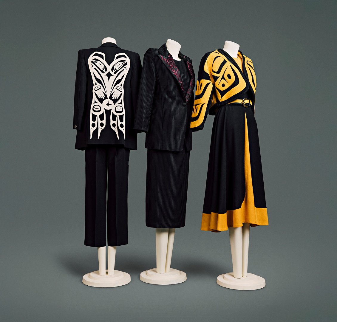 Une collection de trois tailleurs contemporains en laine pour femme. Le premier se compose d’une veste noire ornée d’un motif Haïda blanc dans le dos et d’un pantalon noir arrivant à mi-mollet. Le deuxième est en tissu noir à fines rayures, avec un motif Haïda rouge sur le revers de la veste. Il est accompagné d’un chemisier assorti et d’une jupe arrivant juste sous le genou. Le troisième se compose d’une veste noire courte, façon cape, à motif Haïda jaune vif, portée sur un chemisier jaune et une jupe jaune et noire arrivant juste sous le genou. Une ceinture en cuir noir et jaune à boucle simple en forme de croissant complète l’ensemble.