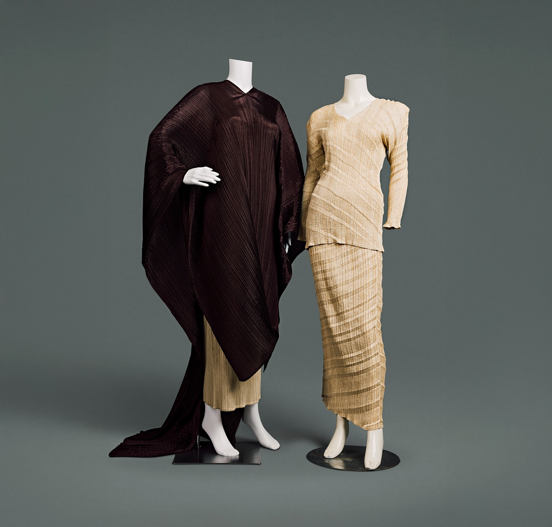 Deux ensembles contemporains en polyester. Le premier se compose d’un chemisier beige ajusté et d’une jupe longue dont le tissu est froissé verticalement et rayé horizontalement en diagonale. Le second se compose d’une jupe similaire, en grande partie recouverte d’une tunique marron foncé qui couvre les bras et le corps, descend sous les genoux grâce à une découpe en V sur le devant et se prolonge jusqu’au sol comme une longue cape à l’arrière.