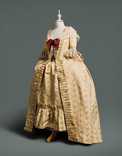 Une robe couleur crème, ornée d’un empiècement fluide au dos du corsage, dévoilant un tissu précieux à motifs de grenades, de roses et autres fleurs, sur un fond de dentelle damassée. Un grand nœud auburn et un châle de dentelle drapent le haut de la robe.