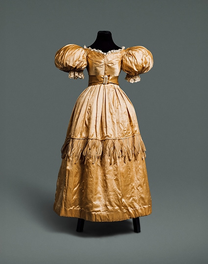 Robe en soie dorée avec une longue jupe ample, des manches bouffantes autour des épaules et une taille fine soulignée par une ceinture de même matière.