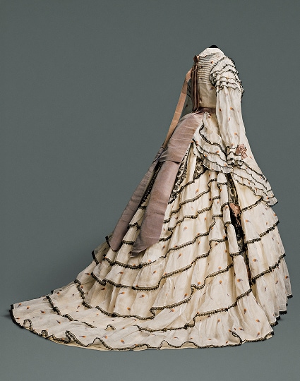 Une longue jupe volumineuse composée de plusieurs couches de coton et de lin à motifs couleur crème, rehaussée de dentelle noire. Une ceinture marron clair descend de la taille, à l’arrière de la robe.