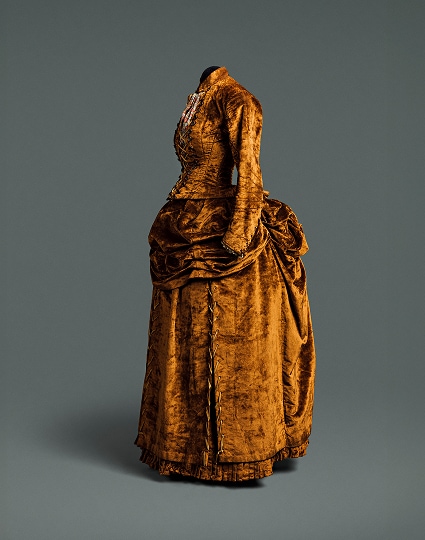 Une robe ocre foncé composée d’un corsage et d’une jupe séparés. La robe est ornée de boutons décoratifs marron qui descendent le long du corsage et sont lacés par un cordon doré.