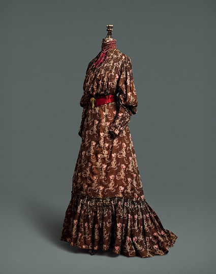 Une robe quatre pièces en coton marron chocolat à motifs de chardons marron clair et roses. La robe est rehaussée d’une ceinture en soie rouge foncé, fermée par une agrafe dorée, et d’un col.