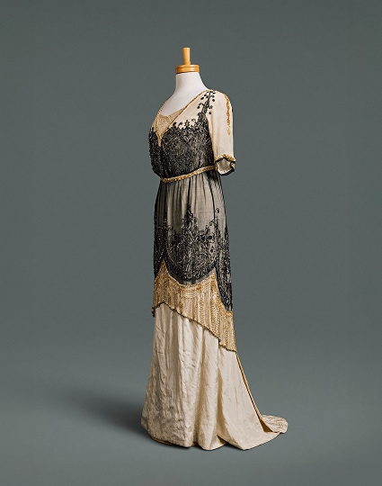 Une longue robe étroite en soie couleur crème, à décolleté plongeant et taille haute, recouverte d’une surjupe tunique ornée de perles noires et dorées.