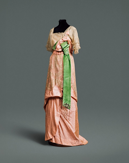 Une robe en soie rose à jupe drapée et large ceinture vert émeraude, recouverte d’un corsage et d’une surjupe en dentelle finement travaillée.