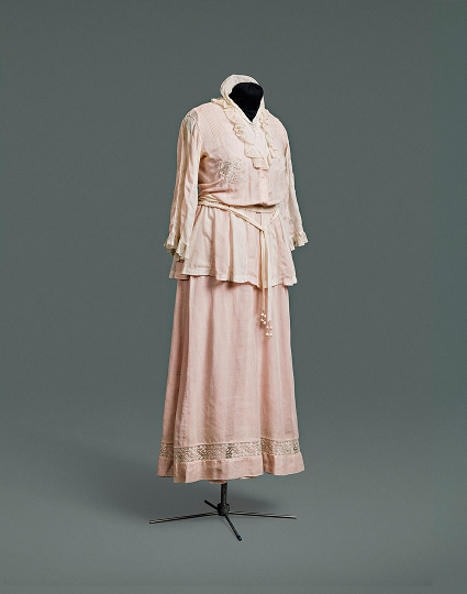 Une simple robe-robe rose en coton, conçue pour ressembler à un tailleur composé d’une veste sur un chemisier, portée avec une jupe assortie.