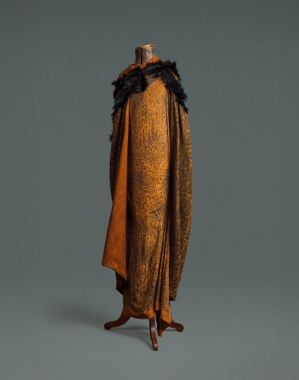 Une longue robe ample en soie auburn ornée d’un motif floral en perles noires. Elle est recouverte d’une cape assortie en tissu plus épais à motifs et d’un boa en plumes noires.