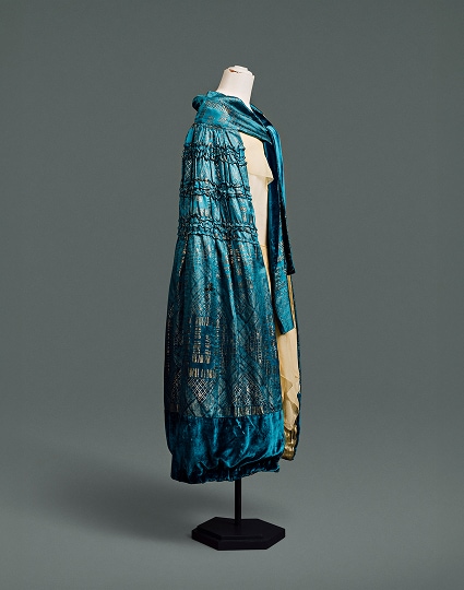 Une cape en soie bleue, ornée d’un motif damassé Art déco aux formes géométriques dorées métallisées, arrive juste en dessous du genou.