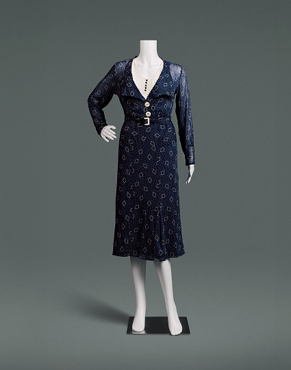 Une robe en coton bleu nuit, cintrée, à taille haute et à jupe arrivant juste sous les genoux. Le tissu est orné de petits pois et d’un motif en losanges.