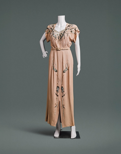Une robe couleur pêche ornée de perles et de fleurs, aux épaules fortement rembourrées et à la jupe ajustée. Une fine ceinture du même tissu souligne la taille haute.