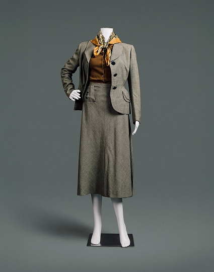 Un tailleur en laine grise anglaise, composé d’une veste et d’une jupe. Ce tailleur présente des épaules tombantes et larges, une blouse boutonnée auburn, une taille fine et une jupe longue et ample. Un foulard à motifs cachemire noué autour du cou apporte la touche finale parfaite.
