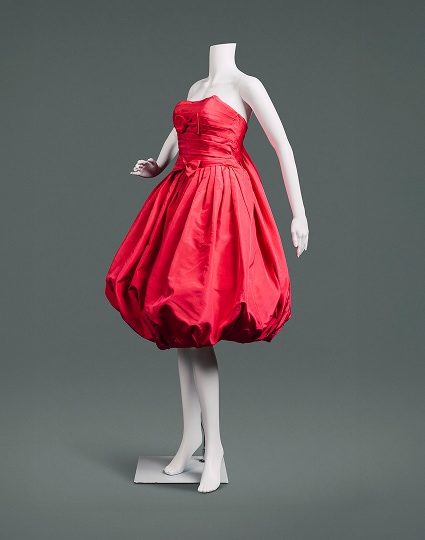 Une robe cocktail rouge cerise sans manches en soie, avec une jupe ballon, un dos sac et des nœuds rouges au niveau de la poitrine et de la taille.