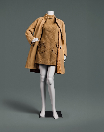 Une robe courte en laine couleur camel, à col roulé, à motif chevrons et poches en biais, accompagnée d’un épais manteau camel qui descend juste au-dessus du genou.