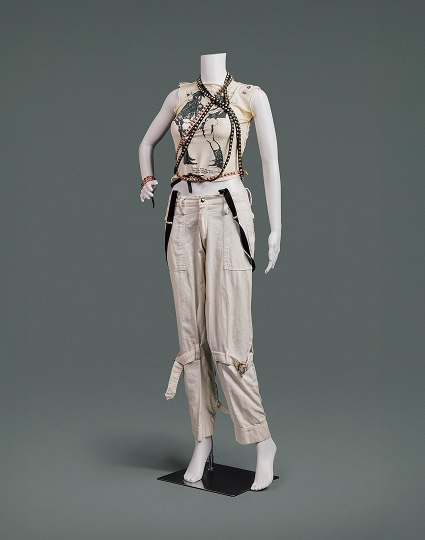 Un t-shirt à manches déchirées orné de l’emblématique imprimé « Two Cowboys » de Vivienne Westwood sur le devant, un pantalon en coton couleur crème avec de grandes poches et des ceintures cousues qui se resserrent autour de chaque jambe au-dessus du genou, et des ceintures en cuir cloutées noires et roses drapées autour du cou par-dessus le t-shirt.