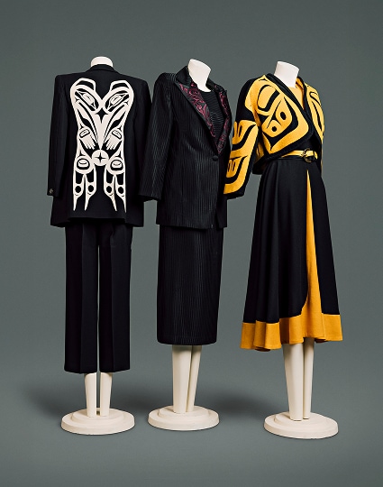 Une collection de trois tailleurs contemporains en laine pour femme. Le premier se compose d’une veste noire ornée d’un motif Haïda blanc dans le dos et d’un pantalon noir arrivant à mi-mollet. Le deuxième est en tissu noir à fines rayures, avec un motif Haïda rouge sur le revers de la veste. Il est accompagné d’un chemisier assorti et d’une jupe arrivant juste sous le genou. Le troisième se compose d’une veste noire courte, façon cape, à motif Haïda jaune vif, portée sur un chemisier jaune et une jupe jaune et noire arrivant juste sous le genou. Une ceinture en cuir noir et jaune à boucle simple en forme de croissant complète l’ensemble.