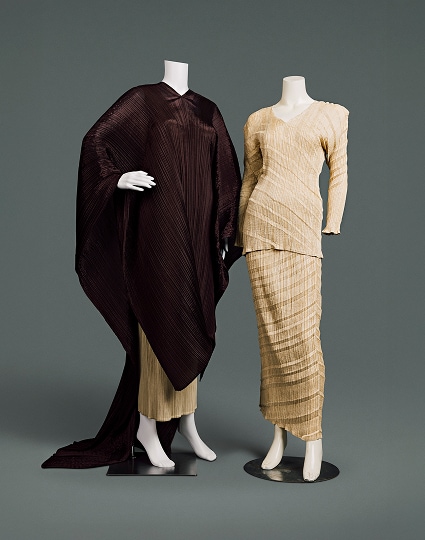 Deux ensembles contemporains en polyester. Le premier se compose d’un chemisier beige ajusté et d’une jupe longue dont le tissu est froissé verticalement et rayé horizontalement en diagonale. Le second se compose d’une jupe similaire, en grande partie recouverte d’une tunique marron foncé qui couvre les bras et le corps, descend sous les genoux grâce à une découpe en V sur le devant et se prolonge jusqu’au sol comme une longue cape à l’arrière.