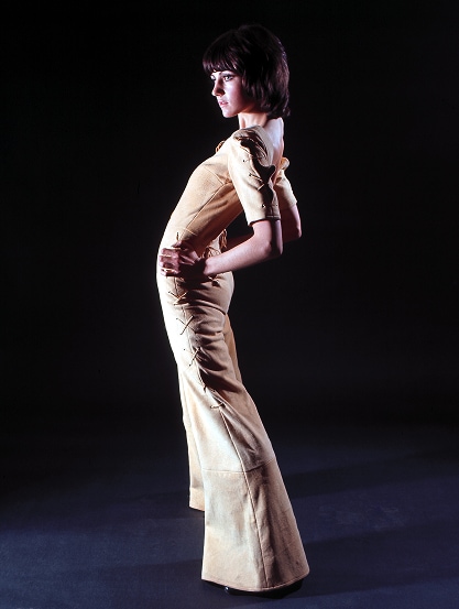 Un mannequin des années 1970, vêtue d’un tailleur-pantalon une pièce, pose le dos cambré et les mains sur les hanches.