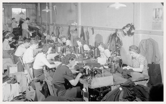 Un grand groupe de femmes sont assises de part et d’autre d’une longue table, assemblant des vêtements à l’aide de machines à coudre.