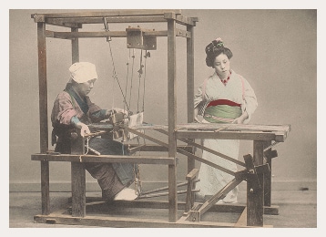 Une Japonaise est assise sur un métier à tisser tandis qu’une autre y alimente la machine en fil. Toutes deux portent un kimono traditionnel.