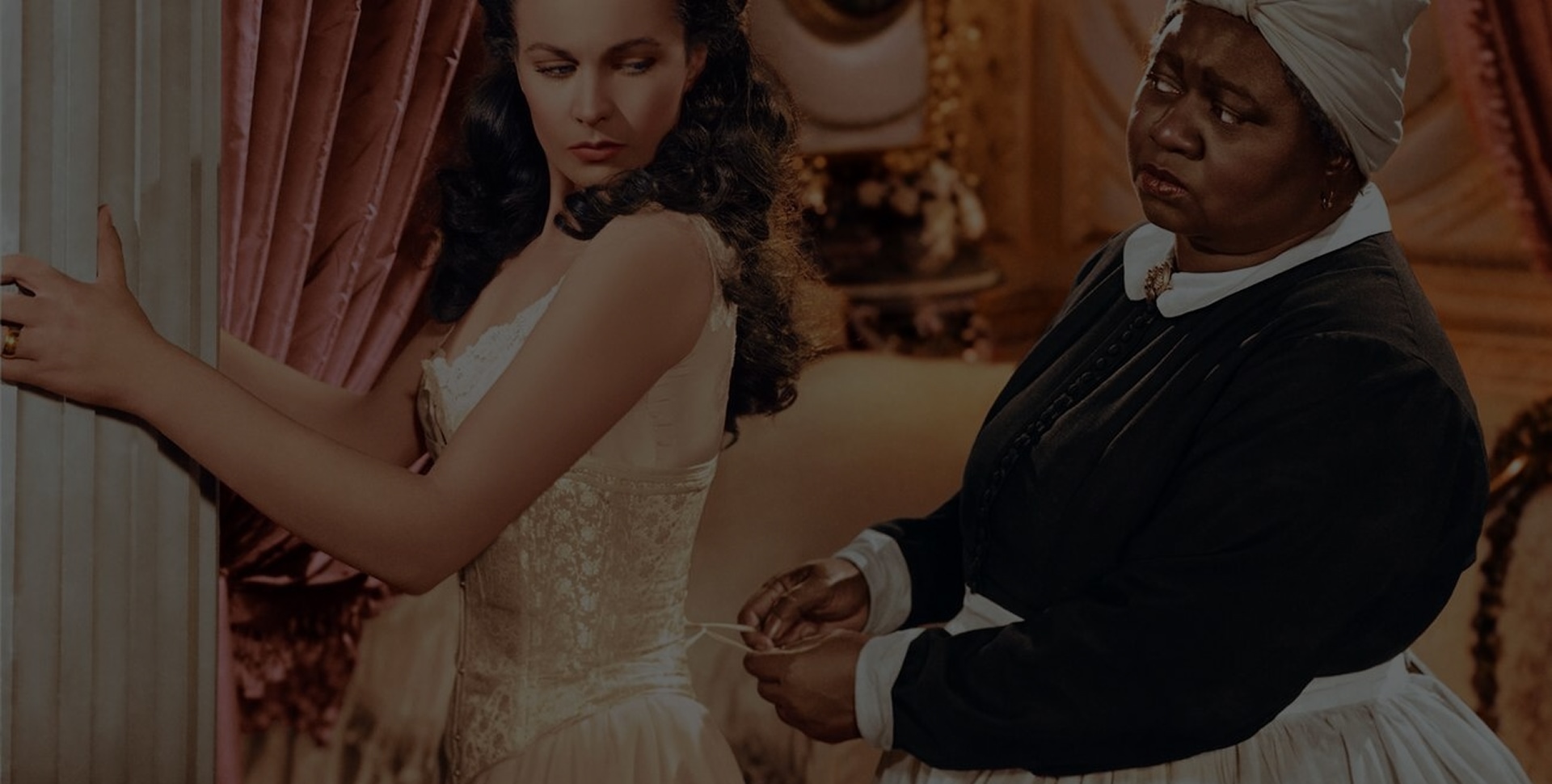 Une photo de plateau du film Gone with the Wind montre Mammy en train de resserrer le corset de Scarlett tandis que celle-ci s’appuie contre une colonne.