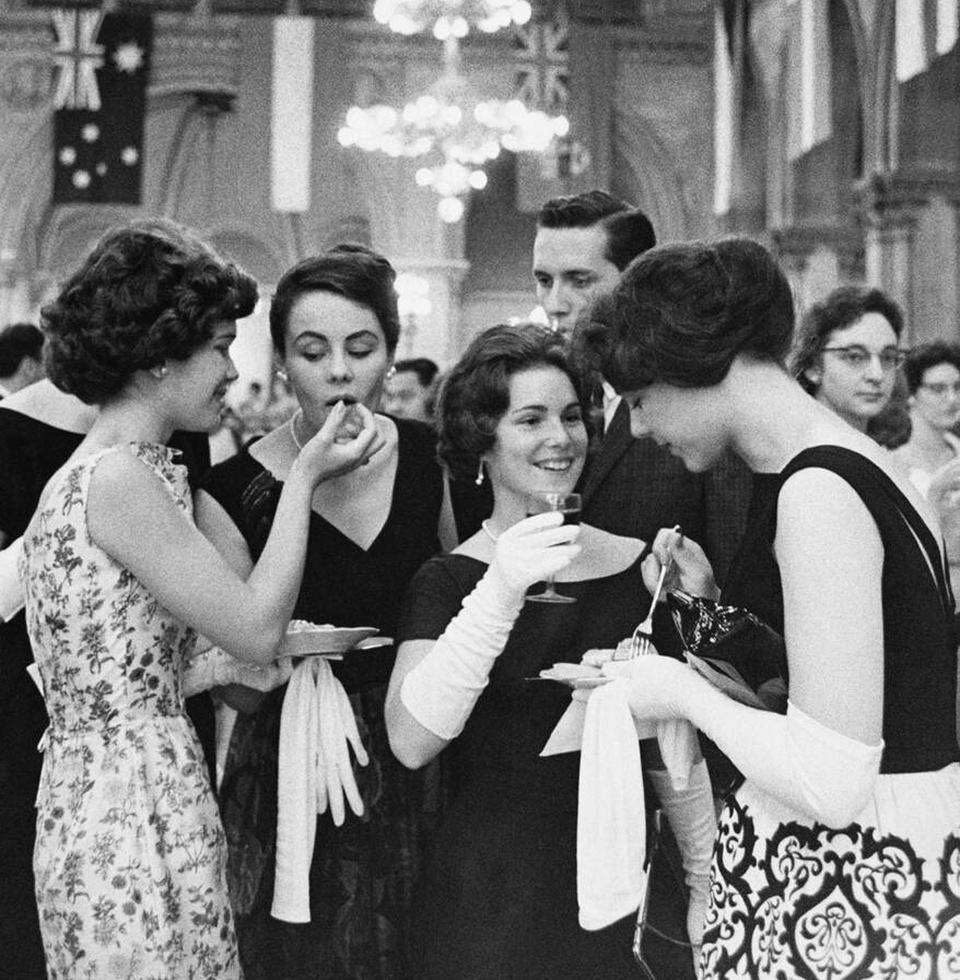 Quatre femmes, portant de longs gants blancs, mangent du gâteau et boivent du vin lors d’une soirée cocktail huppée.