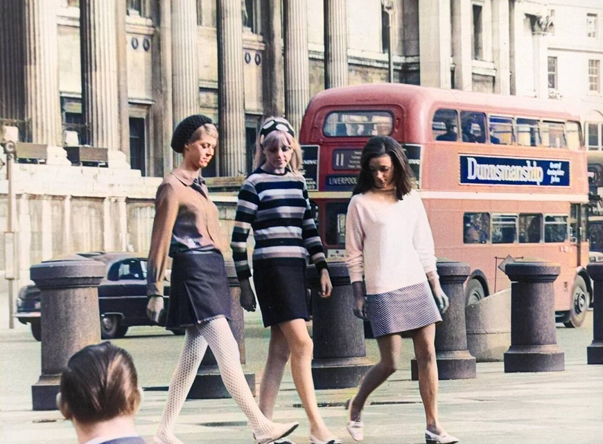 Trois jeunes femmes en minijupes traversent Trafalgar Square, un bus à impériale en arrière-plan.