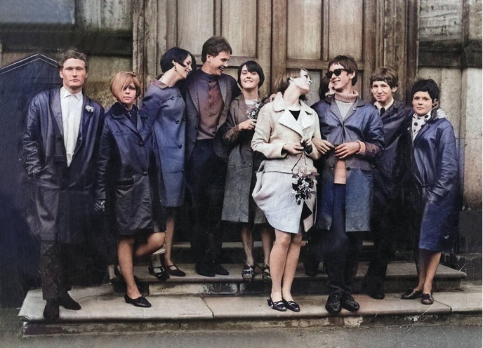 Un groupe de couples modernes et élégants, vêtus de longs manteaux, se tient sur les marches de l’entrée d’un vieux bâtiment.
