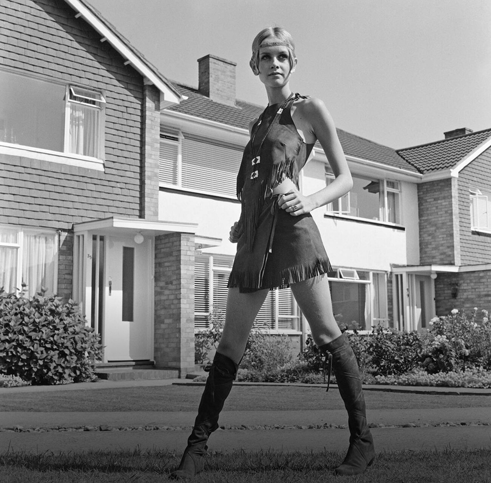 Twiggy, vêtue d’une mini-robe en cuir à franges et de bottes assorties, prend la pose devant des maisons de banlieue.