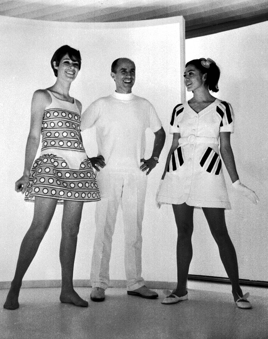 Le créateur de mode André Courrèges est entouré de deux mannequins portant des robes courtes.