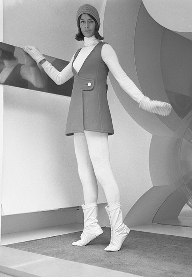 Le mannequin porte une mini-robe sans manches en laine vert vif, un bonnet en laine orange, des moufles blanches et des bottines à boutons.