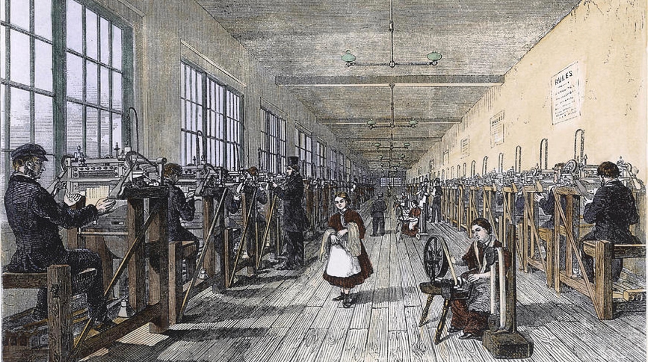 Illustration d’une longue usine de bas victorienne où l’on voit deux rangées d’hommes actionnant des machines tandis que des femmes filent la fibre entre eux.