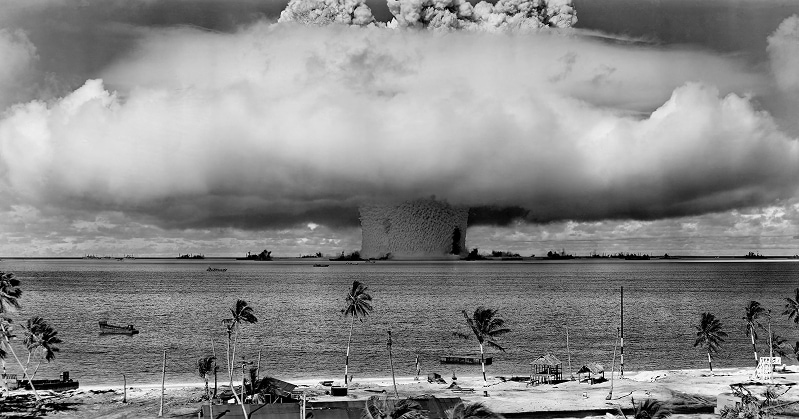 Un gigantesque champignon atomique se forme au-dessus d’un atoll du Pacifique. Des navires entourent l’explosion, tandis qu’une plage et des palmiers sont visibles au premier plan.